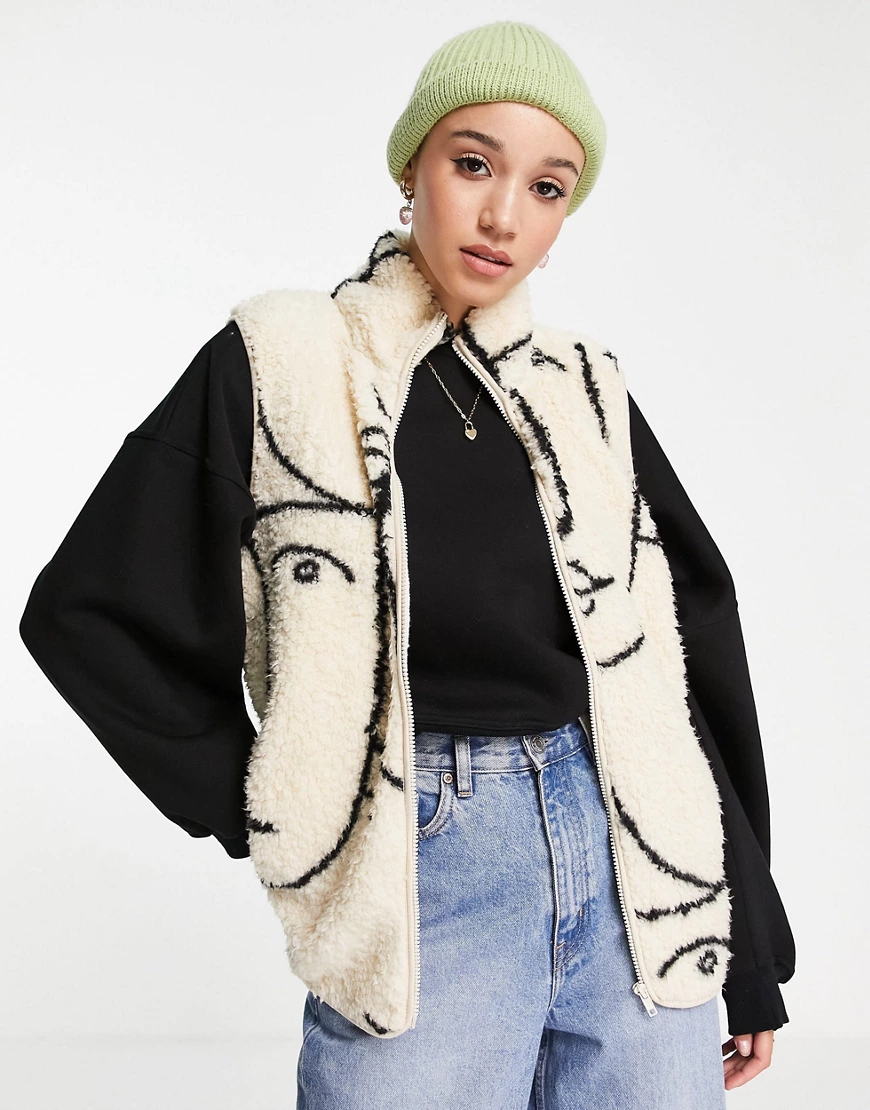 Monki – Weste aus recyceltem Fleece in gebrochenem Beige mit Gesichts-Print günstig online kaufen